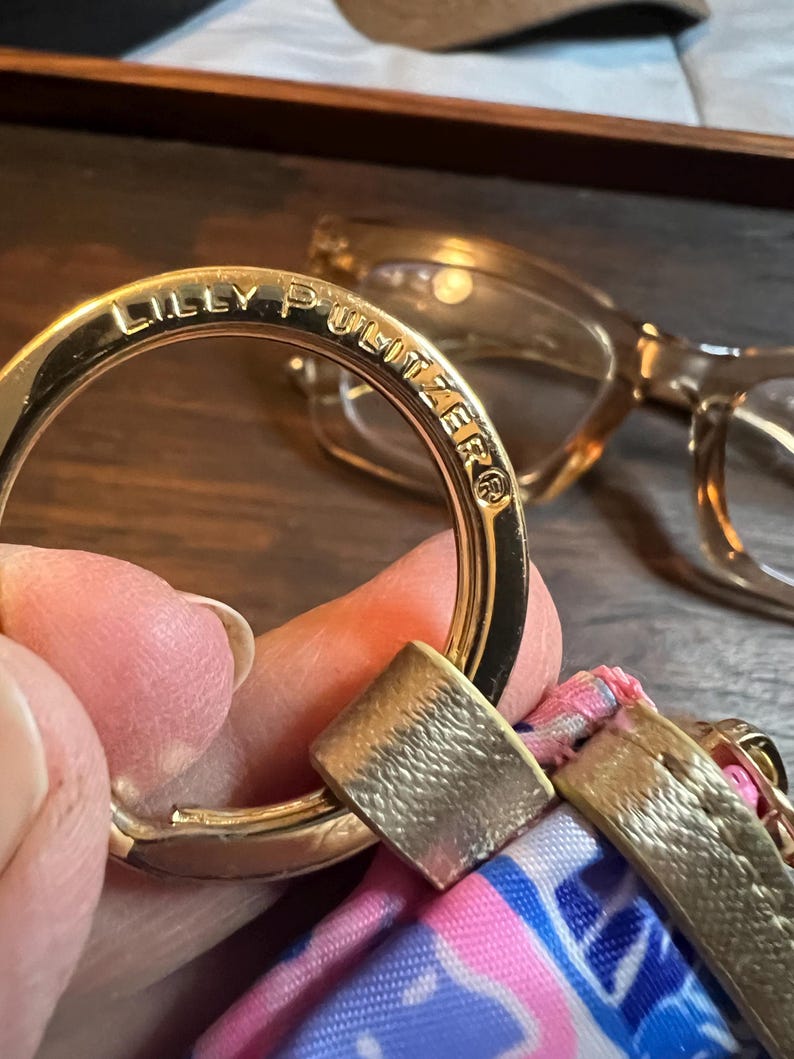 May include: A gold-toned metal keyring featuring the engraved text LILLY PULITZER. The circular ring is attached to a fabric accessory with a pink, blue, and white patterned design, secured by a metallic gold-colored connector piece.