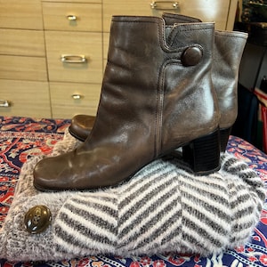 May include: A pair of brown leather ankle boots with a square toe, a block heel, and a single circular button detail on the side, resting on a folded gray and white chevron patterned knit garment with a large button.