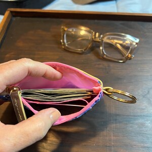 May include: A small zippered pouch with a bright pink interior lining, containing several folded paper bills. The pouch features a gold-toned metal key ring attached to the side and a gold zipper pull. The exterior has a blue and white patterned design.