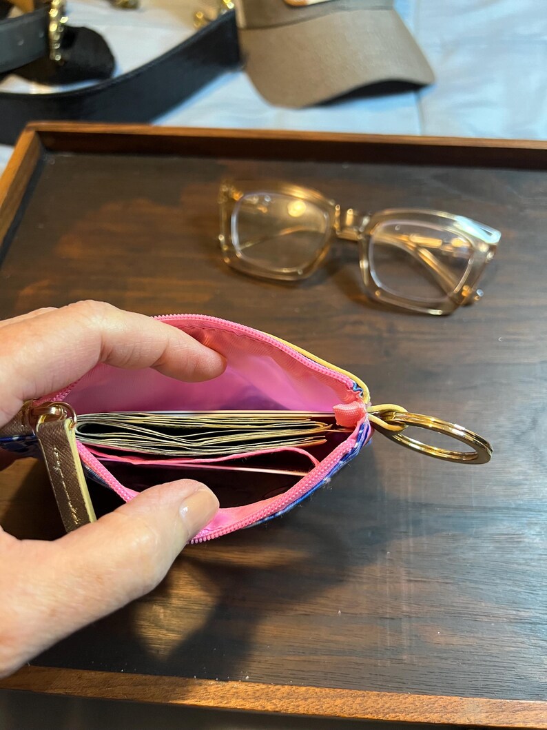 May include: A small zippered pouch with a bright pink interior lining, containing several folded paper bills. The pouch features a gold-toned metal key ring attached to the side and a gold zipper pull. The exterior has a blue and white patterned design.