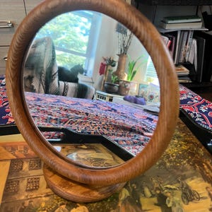 May include: A round, wooden-framed mirror with a light brown finish. The mirror is mounted on a circular wooden base. The reflection shows a room with a window and a bed.