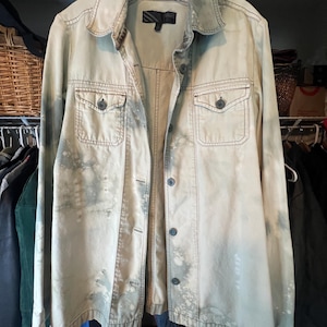 May include: A long-sleeve button-down shirt with a light beige base and a mottled, bleached-effect pattern in shades of gray and white. The shirt features a pointed collar, two chest pockets with button flaps, and a Vans brand label at the neck.