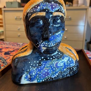May include: A ceramic bust with a dark blue face and a galaxy-like pattern of white dots and blue hues. The hair is a golden-orange colour. The bust is a unique piece of art.