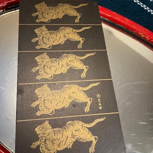 May include: A set of four black coasters with a gold tiger design. The tigers are depicted in a running pose. The coasters are rectangular and appear to be made of a durable material.