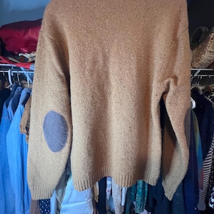 Könnte beinhalten: Ein beigefarbener Pullover mit Ellbogenpatches. Der Pullover ist aus einem strukturierten Strickstoff gefertigt und hat einen Rundhalsausschnitt. Die Ellbogenpatches sind in einem dunkleren Braunton gehalten. Der Pullover hängt an einem Holzbügel.
