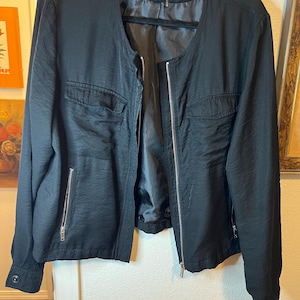 May include: A black DKNY jacket featuring a collarless round neckline, a full-length silver-tone front zipper, and long sleeves with buttoned cuffs. The garment includes two horizontal zippered pockets at the waist and two flap pockets on the chest.