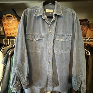 May include: A long-sleeve blue corduroy button-down shirt featuring a classic collar, two chest pockets with button closures, and a front button placket. The shirt has a textured fabric finish and includes a label at the inner neck area.