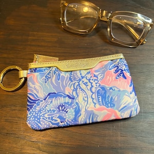 May include: A rectangular zippered pouch featuring a blue, pink, and white floral pattern with a gold metallic trim and a gold key ring attached to the side, positioned below a pair of translucent amber-colored eyeglasses with thick frames.
