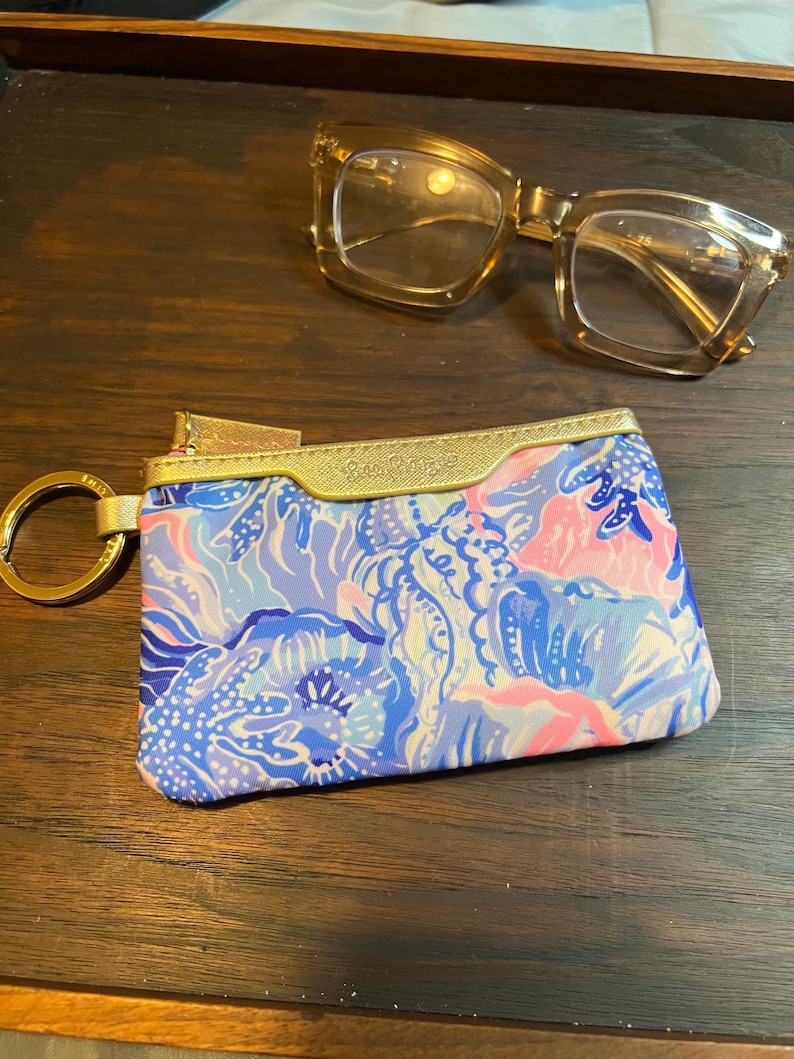 May include: A rectangular zippered pouch featuring a blue, pink, and white floral pattern with a gold metallic trim and a gold key ring attached to the side, positioned below a pair of translucent amber-colored eyeglasses with thick frames.