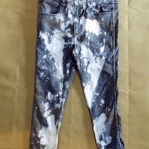 May include: Dark wash denim jeans with white bleach splatter design. The jeans have a high waist and a straight leg cut. The Wrangler brand label is visible on the waistband. The jeans are distressed at the hem.