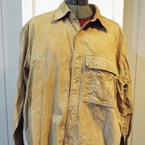 May include: A long-sleeved, tan button-down shirt with a single chest pocket. The shirt has a slightly faded look and is made of a lightweight fabric.