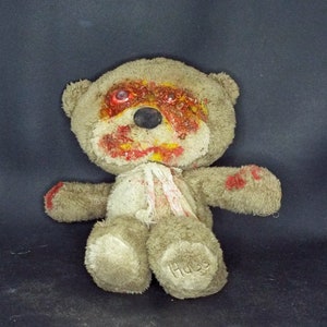 Pode incluir: Um urso de peluche marrom desgastado com respingos de tinta vermelha e amarela no rosto. O urso tem olhos vermelhos e um nariz preto. O urso está usando uma fita branca ao redor do pescoço. O urso está etiquetado com "Hugs" na parte inferior do seu pé.