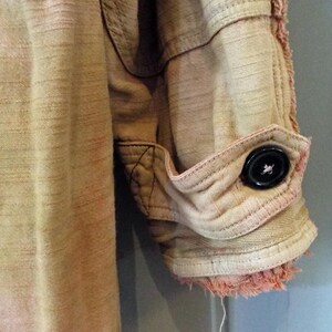 May include: A close-up of a light brown, pink, and white cotton shirt with a frayed hem and a single black button on the cuff.