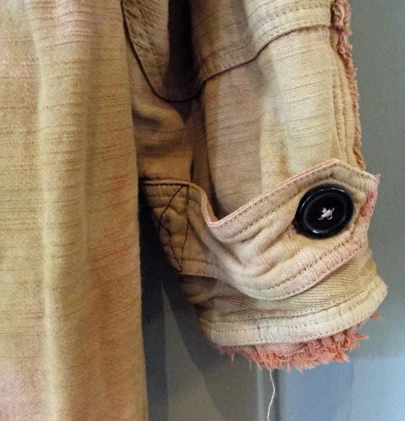 May include: A close-up of a light brown, pink, and white cotton shirt with a frayed hem and a single black button on the cuff.