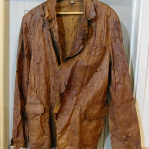 Op de afbeelding: Een bruin leren jack met een verweerde look. Het jack heeft een knoopsluiting en twee zakken. Het leer is versleten en heeft veel gaten.