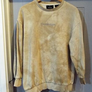 May include: A beige sweatshirt with a tie-dye pattern and black trim on the sleeves. The sweatshirt has the text "CALVIN KLEIN" embroidered on the front.