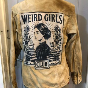 Peut inclure: Veste beige avec un grand motif au dos. Le motif indique "WEIRD GIRLS CLUB" et présente une illustration en noir et blanc d'une femme avec un chignon, entourée de fleurs, de livres et d'une tasse fumante. La veste a un devant boutonné et un col.