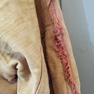 May include: A close-up of a beige fabric with a pink fringe detail. The fabric has a textured, woven appearance.