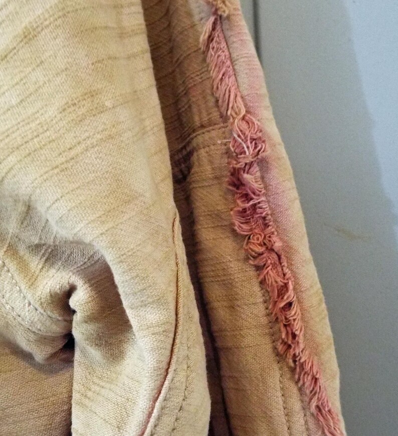 May include: A close-up of a beige fabric with a pink fringe detail. The fabric has a textured, woven appearance.