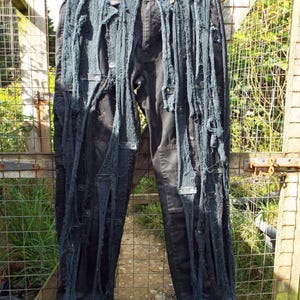 Puede incluir: Vaqueros negros de denim con múltiples tiras largas de tela que cuelgan de las piernas. Los vaqueros están rotos y desgastados.