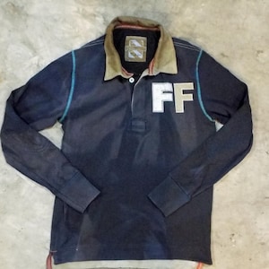 May include: Navy blue long-sleeve polo shirt with a contrasting tan collar and the letters "FF" in white on the chest. The shirt has a button-up placket and teal stitching accents. The cuffs and hem have red accents.