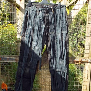 Puede incluir: Pantalón vaquero negro con detalles desgastados y detalles metálicos. El pantalón está colgado de una percha contra una valla de madera.