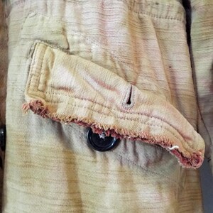 May include: Close-up of a brown and tan striped fabric with a frayed pocket and a black button. The fabric has a worn and faded appearance.