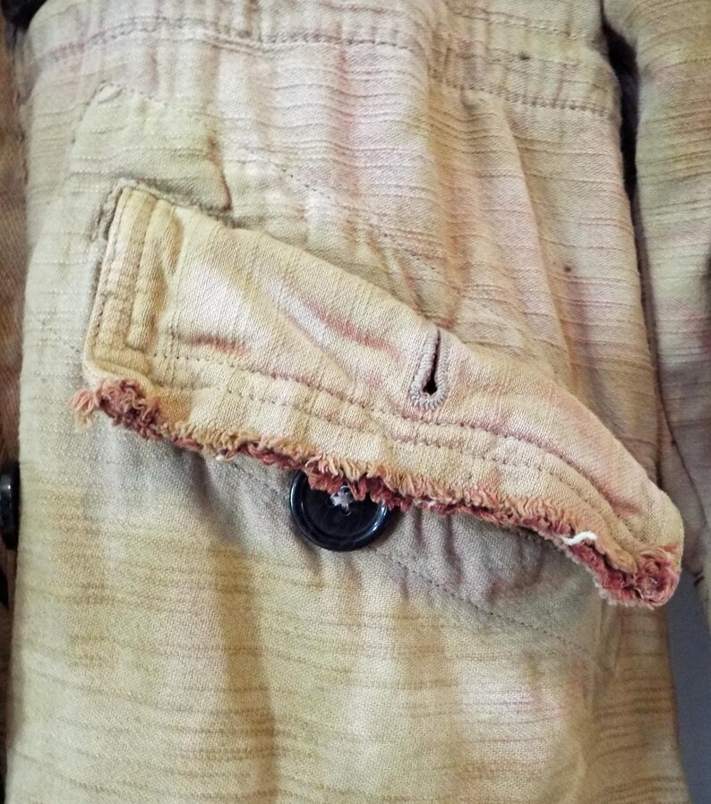 May include: Close-up of a brown and tan striped fabric with a frayed pocket and a black button. The fabric has a worn and faded appearance.