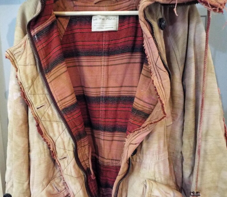 May include: A cream-colored jacket with a red and black plaid lining. The jacket has a zipper closure and a hood. The label on the jacket reads "We The Free".