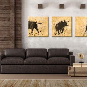 May include: Three framed canvas prints of black bulls on a beige background with brown splatters of paint. The prints are hanging on a white wall above a brown leather sofa.
