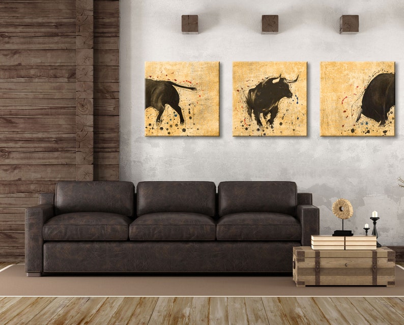 May include: Three framed canvas prints of black bulls on a beige background with brown splatters of paint. The prints are hanging on a white wall above a brown leather sofa.