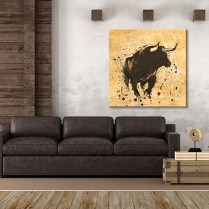 May include: A black and white abstract painting of a bull on a yellow background with splashes of color. The painting is framed and hanging on a wall above a brown leather sofa.
