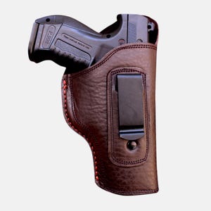 May include: A dark brown leather holster with a black handgun inside. The holster has red stitching and a black metal clip. The handgun has the text "P.998" and "Read Manual Before Use".