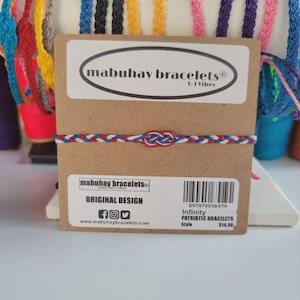 May include: A patriotic infinity bracelet with red, white, and blue braided cord. The bracelet is displayed on a brown card with the text "mabuhay bracelets" and "ORIGINAL DESIGN". The bracelet is a style of patriotic jewelry.