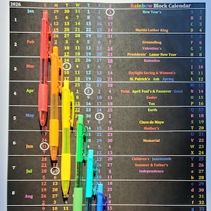 May include: A 2026 calendar with a rainbow-colored block design. The calendar features dates, holidays, and a BestKeptSecretsShop.com website address. Colorful pens are placed along the left and right sides.
