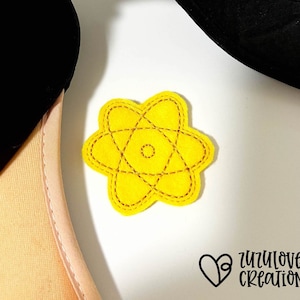 Op de afbeelding: Geel vilt atoomsymbool met zwarte stiksels. Het atoomsymbool is een eenvoudig ontwerp met een cirkel in het midden en vijf ringen eromheen.