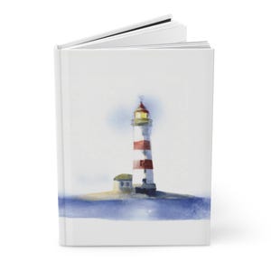 Op de afbeelding: Een hardcover notitieboek met een aquarel schilderij van een vuurtoren. De vuurtoren is wit met rode strepen en een gele top. Het notitieboek is open en toont de lege pagina's. Het beeld heeft een nautisch thema.
