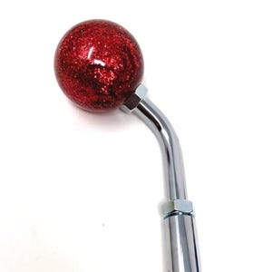 May include: A red glitter ball-shaped gear shift knob with a chrome-plated, curved stem. The knob is a vibrant red with sparkling glitter, and the stem is a shiny, polished metal. The item is set against a white background.