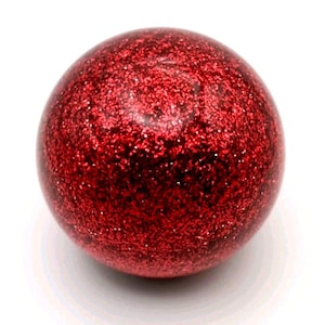 May include: A spherical, red glitter ball. The ball has a shiny, textured surface covered in evenly distributed glitter, creating a sparkling effect. The object is set against a white background.