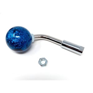 May include: A blue glitter shift knob with a curved, chrome-colored handle and a silver nut. The knob is spherical and has a shiny, textured appearance. The handle is bent and connects to a rectangular end.