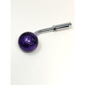 May include: A chrome gear shift knob with a curved arm and a hexagonal end. The round knob is a deep purple with glitter embedded throughout. The knob is designed for a vehicle's gear shift.