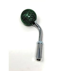 Op de afbeelding: Een groene glitter pookknop met een verchroomde, gebogen handgreep. De pookknop heeft een glanzend, reflecterend oppervlak en is ontworpen voor de versnellingspook van een voertuig.