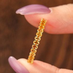 Op de afbeelding: Een delicate gouden ring met een herhalend patroon van kleine, rechthoekige vormen. De ring wordt tussen twee vingers gehouden, wat het slanke ontwerp en de warme, gouden tint benadrukt. De ingewikkelde details van de ring komen uit tegen een vage achtergrond.