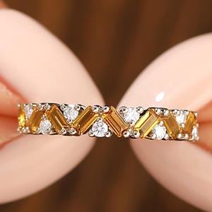 May include: A gold and silver ring with a repeating pattern of yellow and white gemstones. The ring features a series of yellow, rectangular-cut stones alternating with small, round, white stones. The ring is held between two hands.