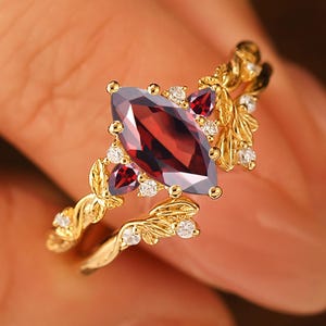 May include: A gold-colored ring featuring a large, marquise-cut, deep red gemstone as the centerpiece. The ring is adorned with smaller red gemstones and clear crystals, with a delicate, leaf-like design along the band.