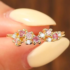 May include: A gold ring featuring a cluster of small, round-cut pink and clear white stones arranged in a delicate, asymmetrical floral-inspired pattern along the top of the thin, polished gold band, creating a sparkling, multi-colored effect.