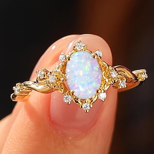 May include: A gold ring featuring a pear-shaped, opalescent gemstone, surrounded by small, clear accent stones. The ring's band has a delicate, twisted design. The ring is held up by a person's hand.