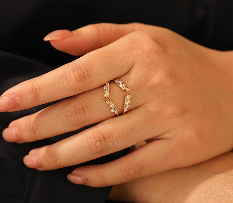 Pu&ograve; includere: Un anello dorato dal design unico, con piccole pietre scintillanti. L'anello ha uno stile aperto e avvolgente con dettagli decorativi a forma di foglie. L'anello &egrave; mostrato su una mano con unghie curate.