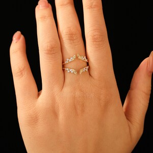 Pu&ograve; includere: Un anello dorato dal design unico, con due fasce curve che si incontrano nella parte superiore e inferiore. Le fasce sono adornate da piccole gemme trasparenti, creando un effetto scintillante. L'anello &egrave; mostrato su una mano su sfondo nero.