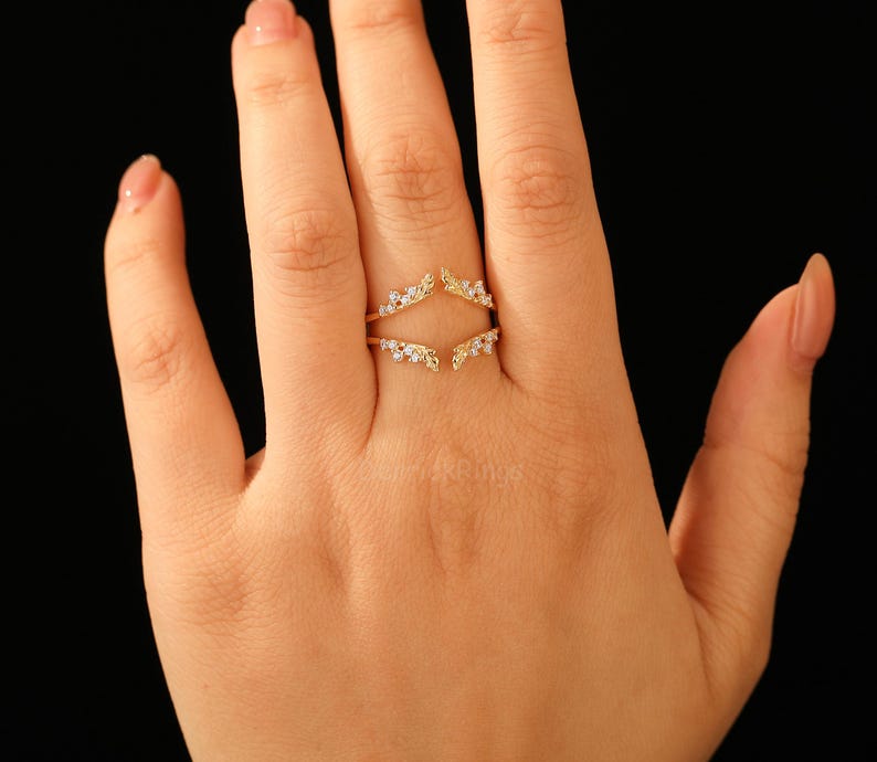 Pu&ograve; includere: Un anello dorato dal design unico, con due fasce curve che si incontrano nella parte superiore e inferiore. Le fasce sono adornate da piccole gemme trasparenti, creando un effetto scintillante. L'anello &egrave; mostrato su una mano su sfondo nero.